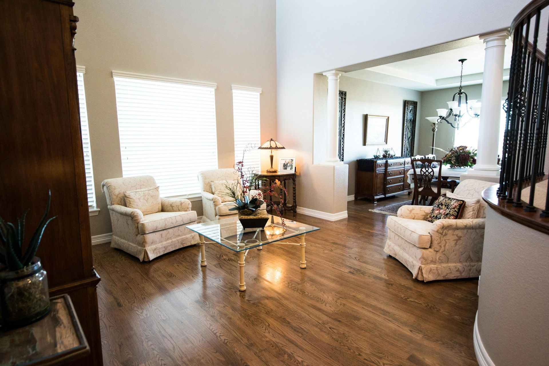 Bright living room with beige chairs, glass coffee table, hardwood floors, and a dining area in the background
