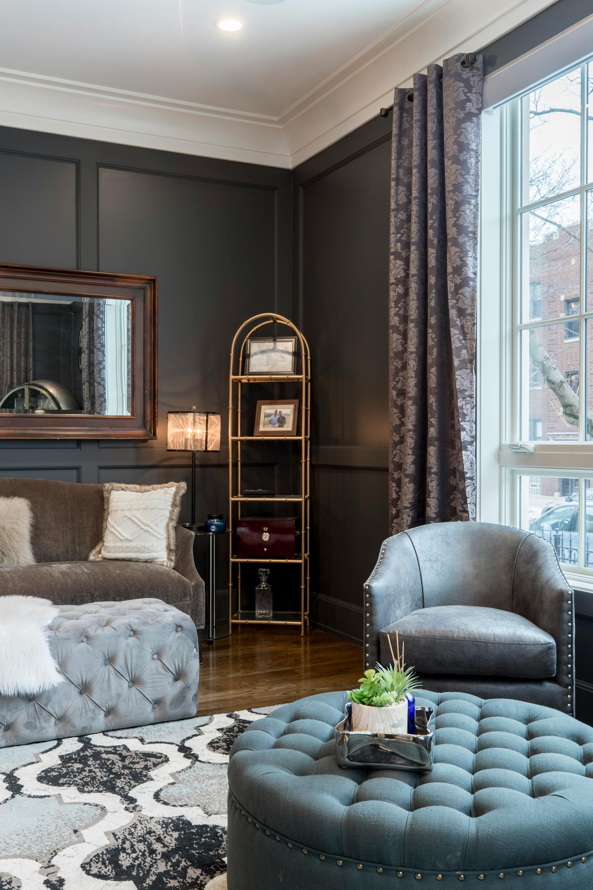 Elegant bedroom with dark paneled walls, gray tufted ottoman, armchair, bed, and tall window with curtains
