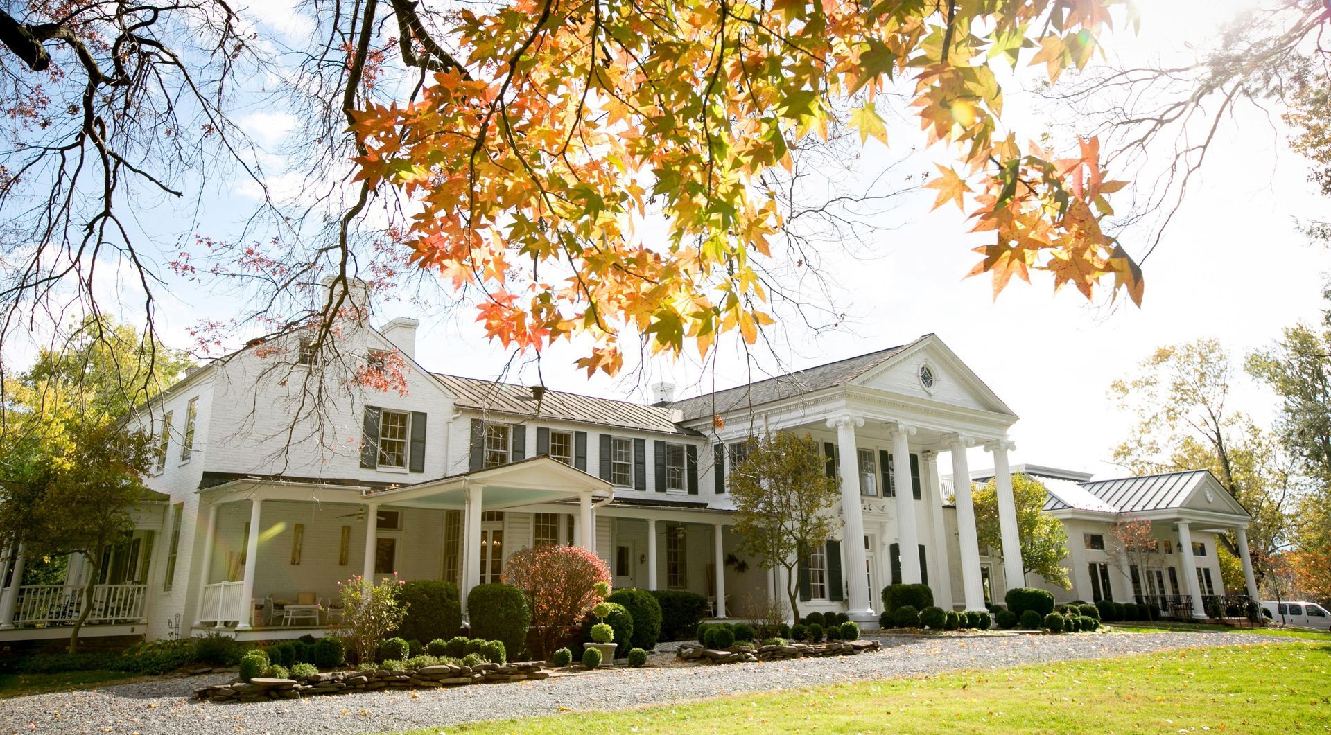 Large white colonial-style house with a columned porch under autumn trees in bright daylight