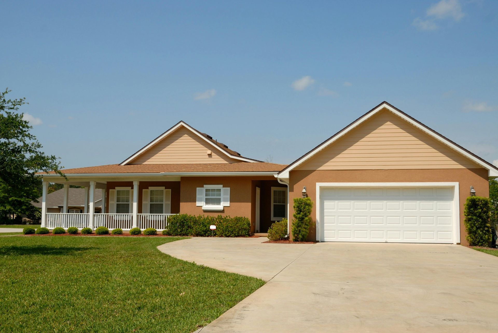 Single-story suburban house with a two-car garage, front porch, and curved driveway under a blue sky