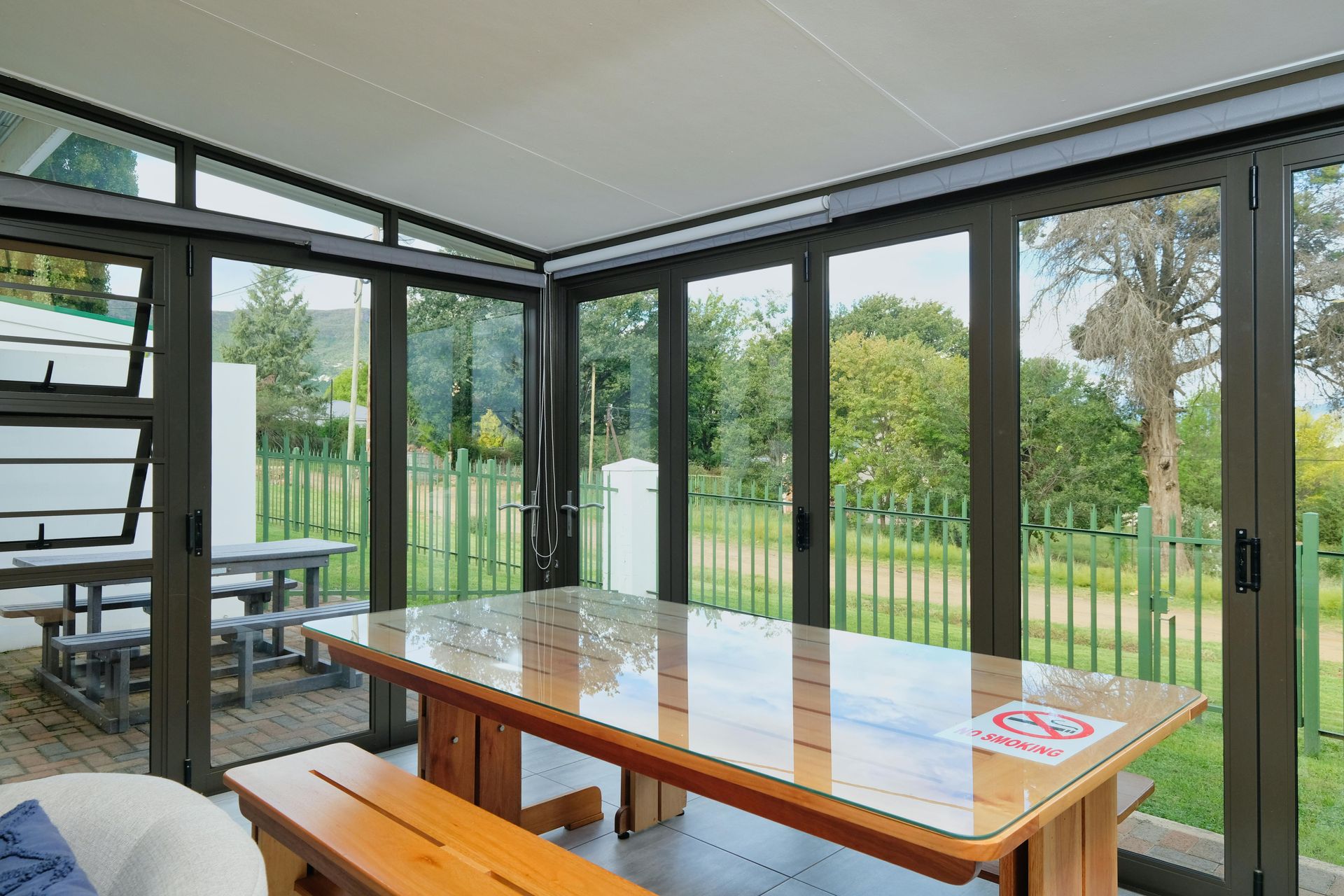 Bright sunroom with glass walls, wooden table and benches, overlooking a green yard and deck.