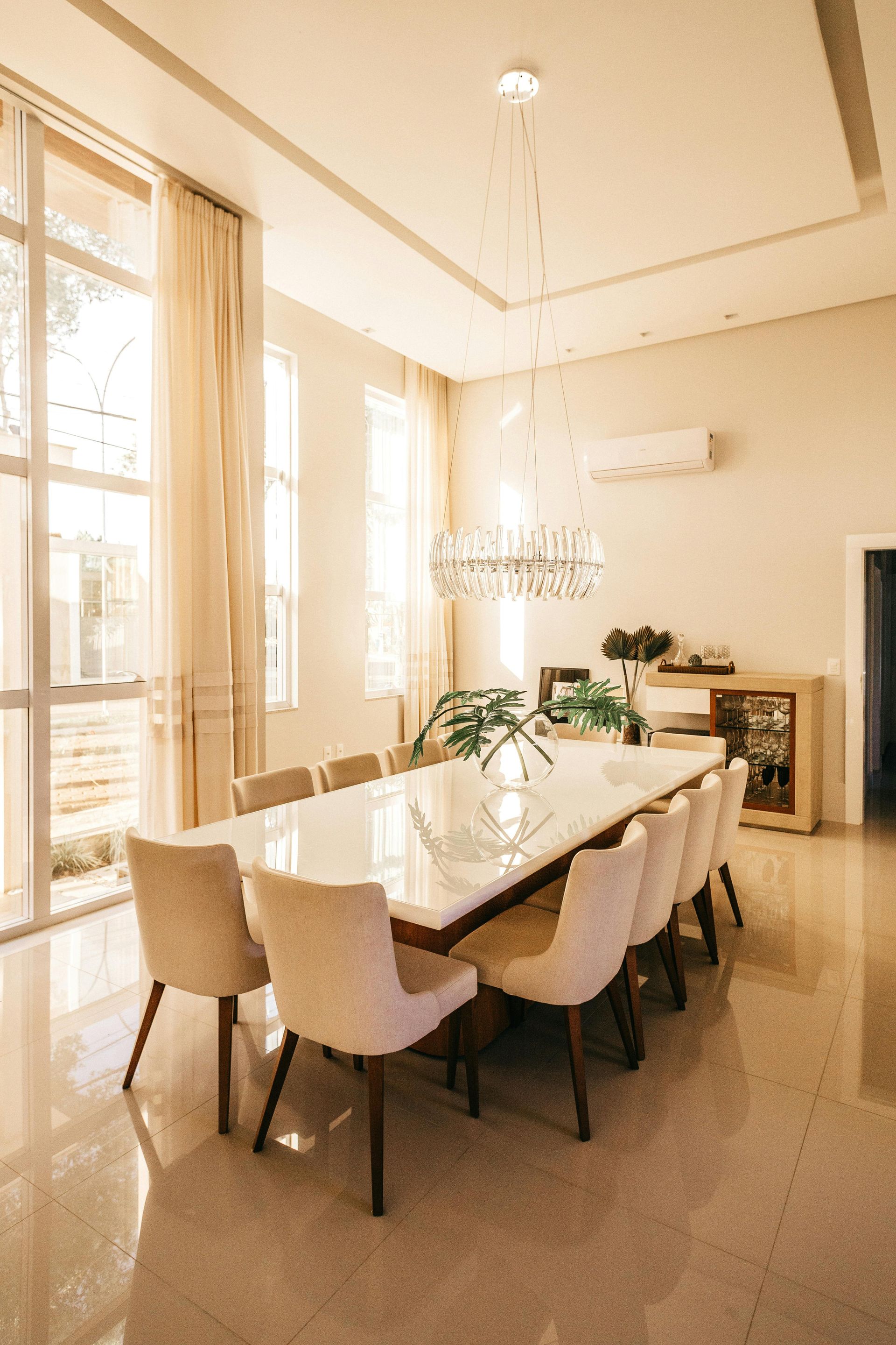 Bright dining room with a long table, white chairs, chandelier, and large windows.