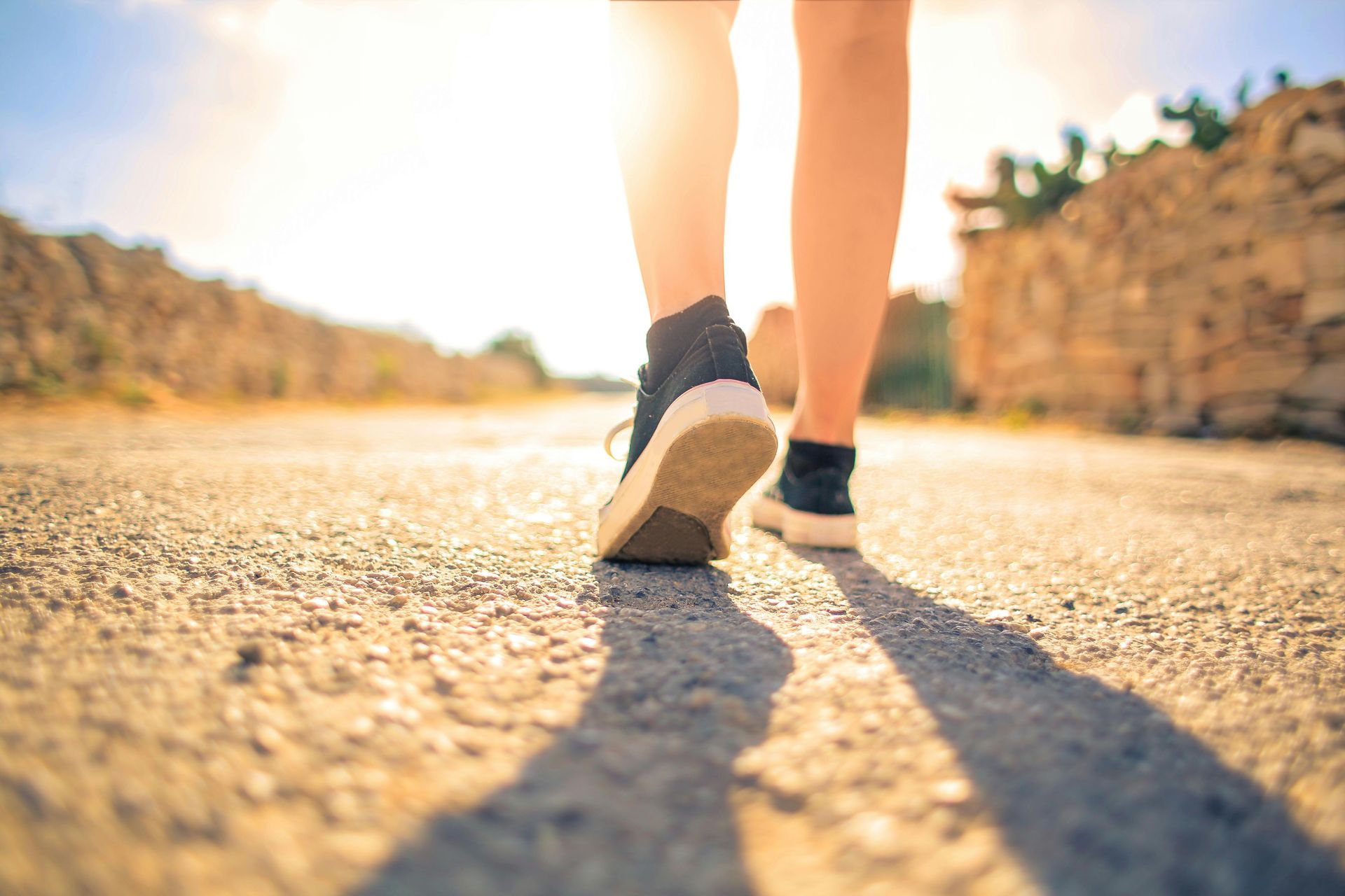 Person walking on a sunlit road, close-up of shoes and legs with long shadows.