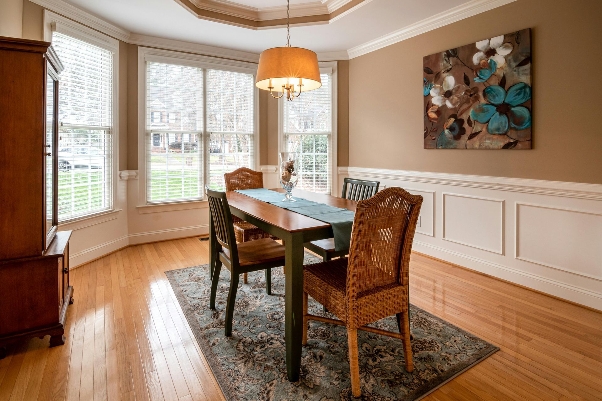 Dining room with wooden table, upholstered chairs, bay windows, and a floral wall painting.