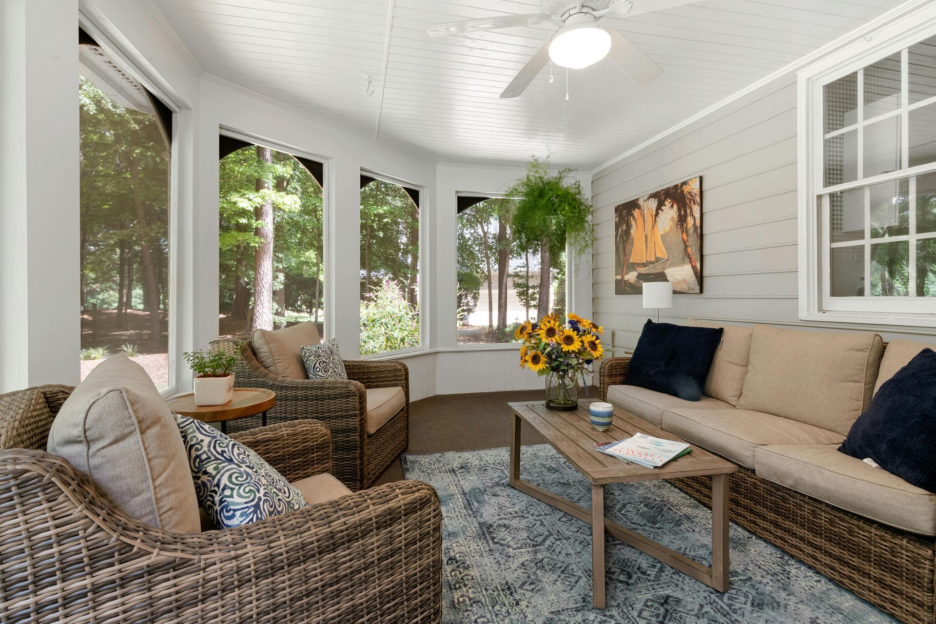 Bright sunroom with wicker seating, beige sofa, blue rug, and sunflowers by large windows