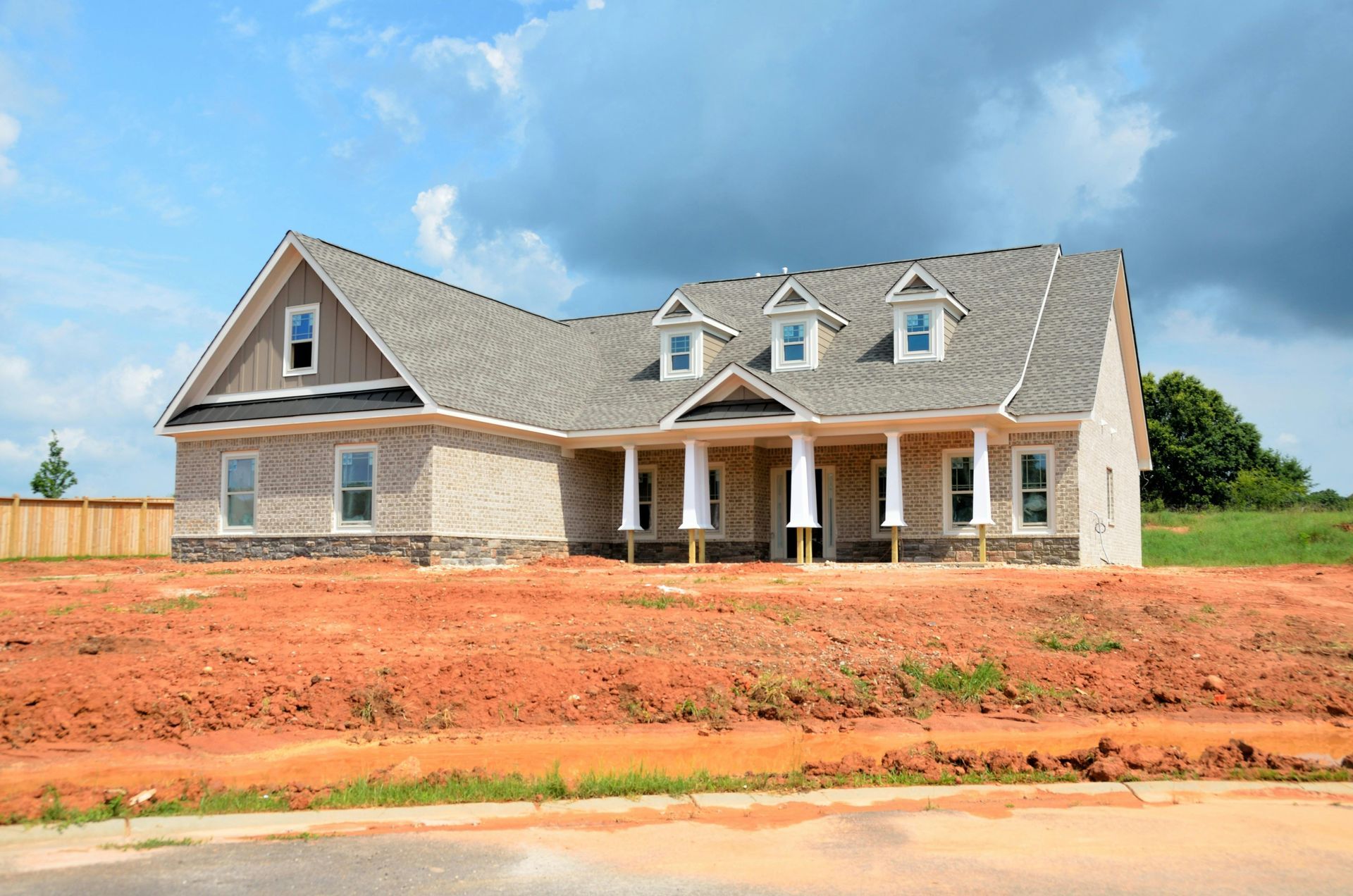 New suburban house with gray roof and brick facade on a dirt lot under a blue sky
