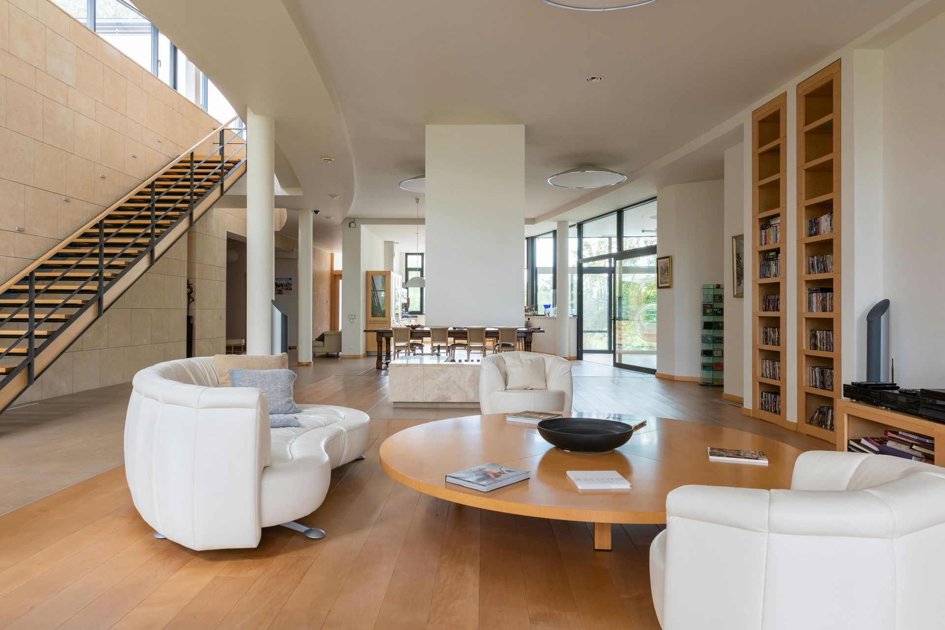 Modern living room with white seating, wood floors, staircase, built-in shelves, and large windows