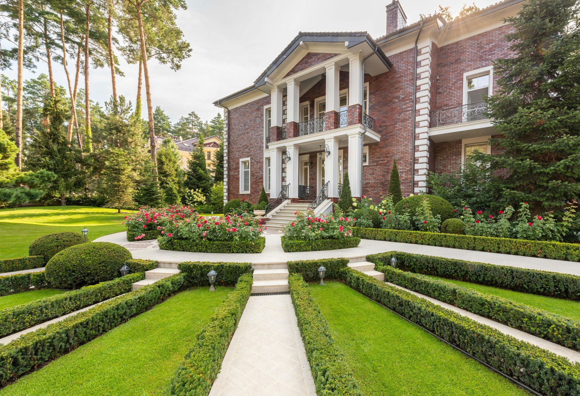 Grand brick mansion with white columns, manicured hedges, and a stone path in a formal garden