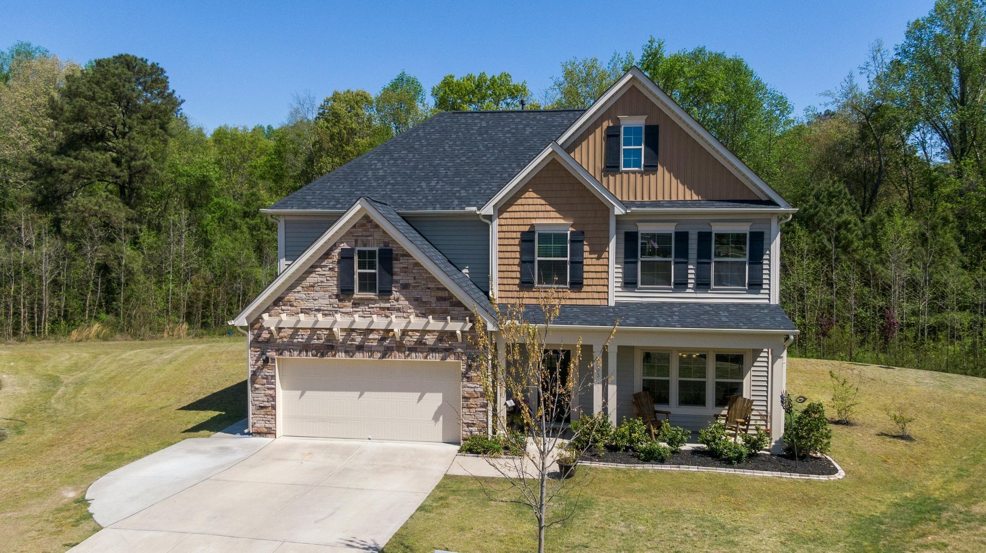 Two-story tan house with attached garage, front porch, and a tree-lined yard under a clear blue sky