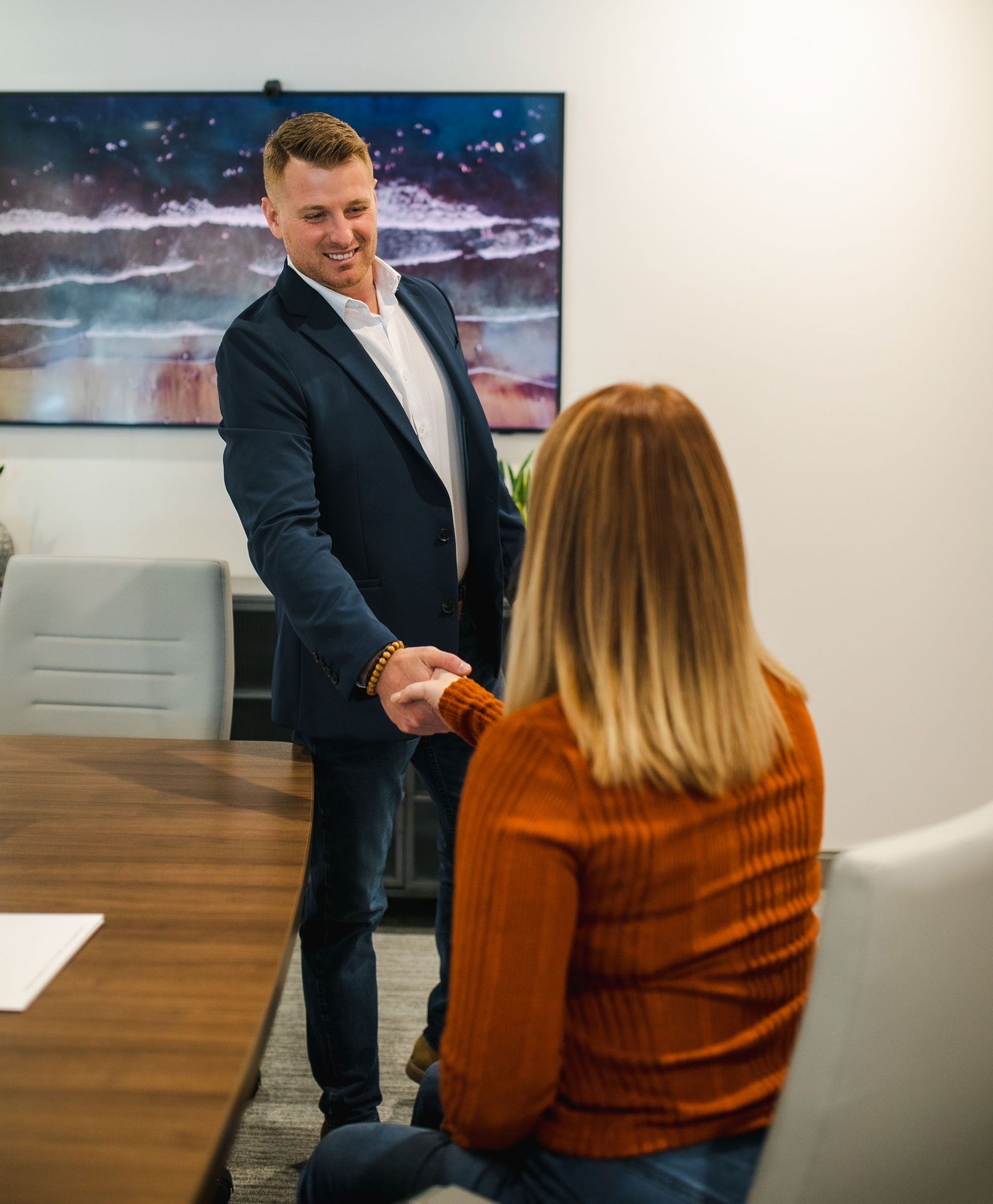 Man shaking hands with a seated woman in a conference room, with a landscape photo on the wall.