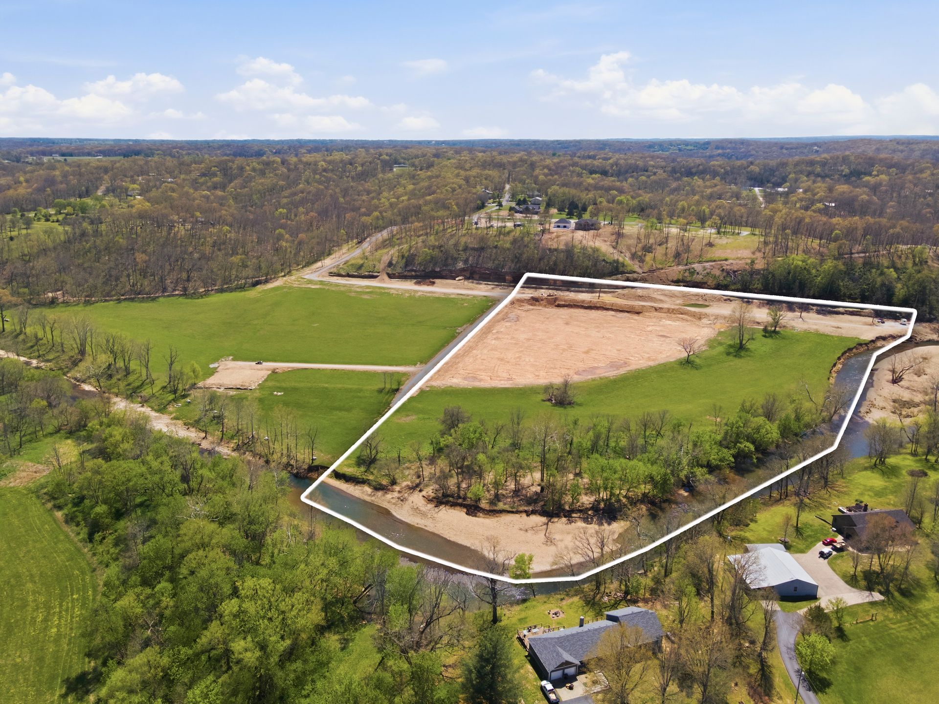 Aerial view of a wooded landscape with a large fenced clearing and a dirt lot near a road.