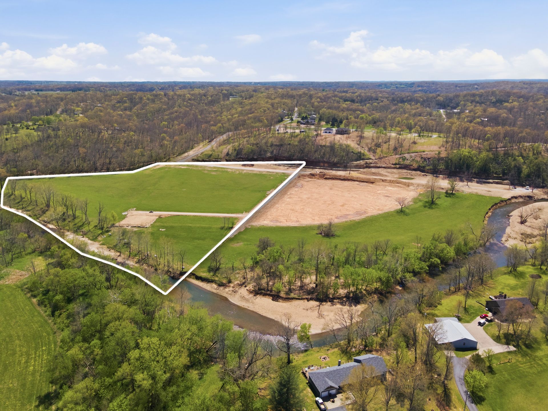 Aerial view of a green field outlined in white beside a tan cleared lot and wooded landscape