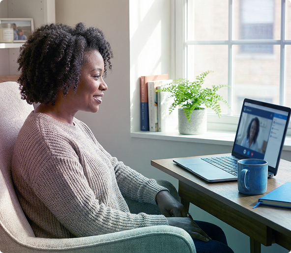 Woman sitting in a chair, smiling at a laptop on a desk beside a blue mug and window plants
