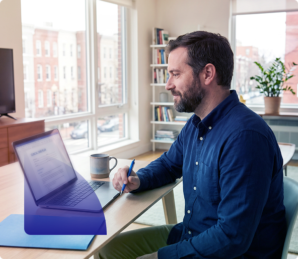 Man smiling while typing on a laptop at a desk with a coffee mug in a bright home office