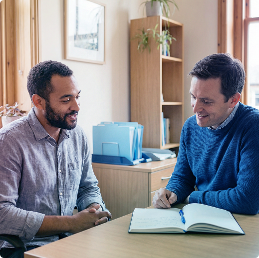 Two men in a bright office talking at a table with an open notebook and folders nearby
