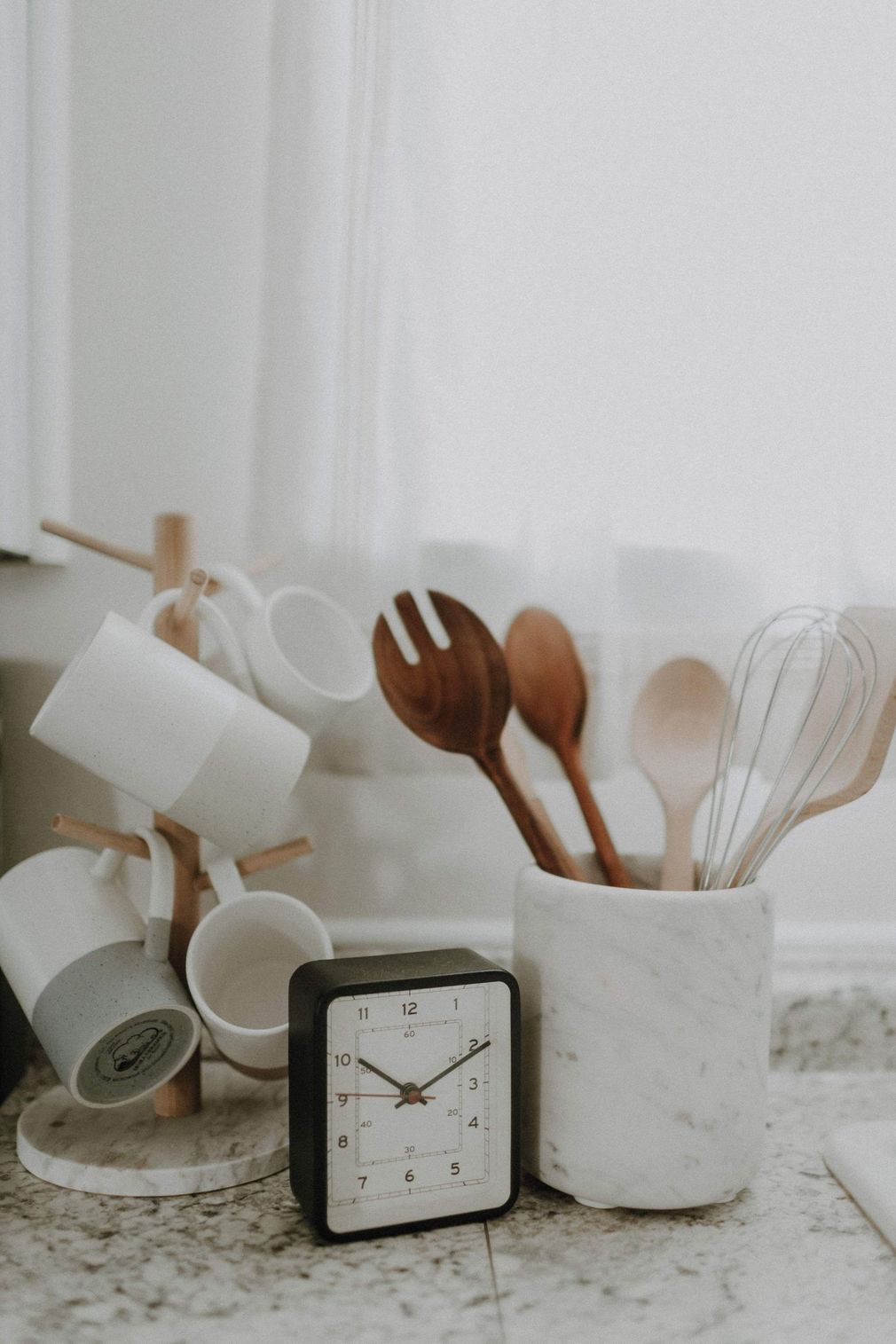 Kitchen utensils in white ceramic holders beside a clock on a light countertop.