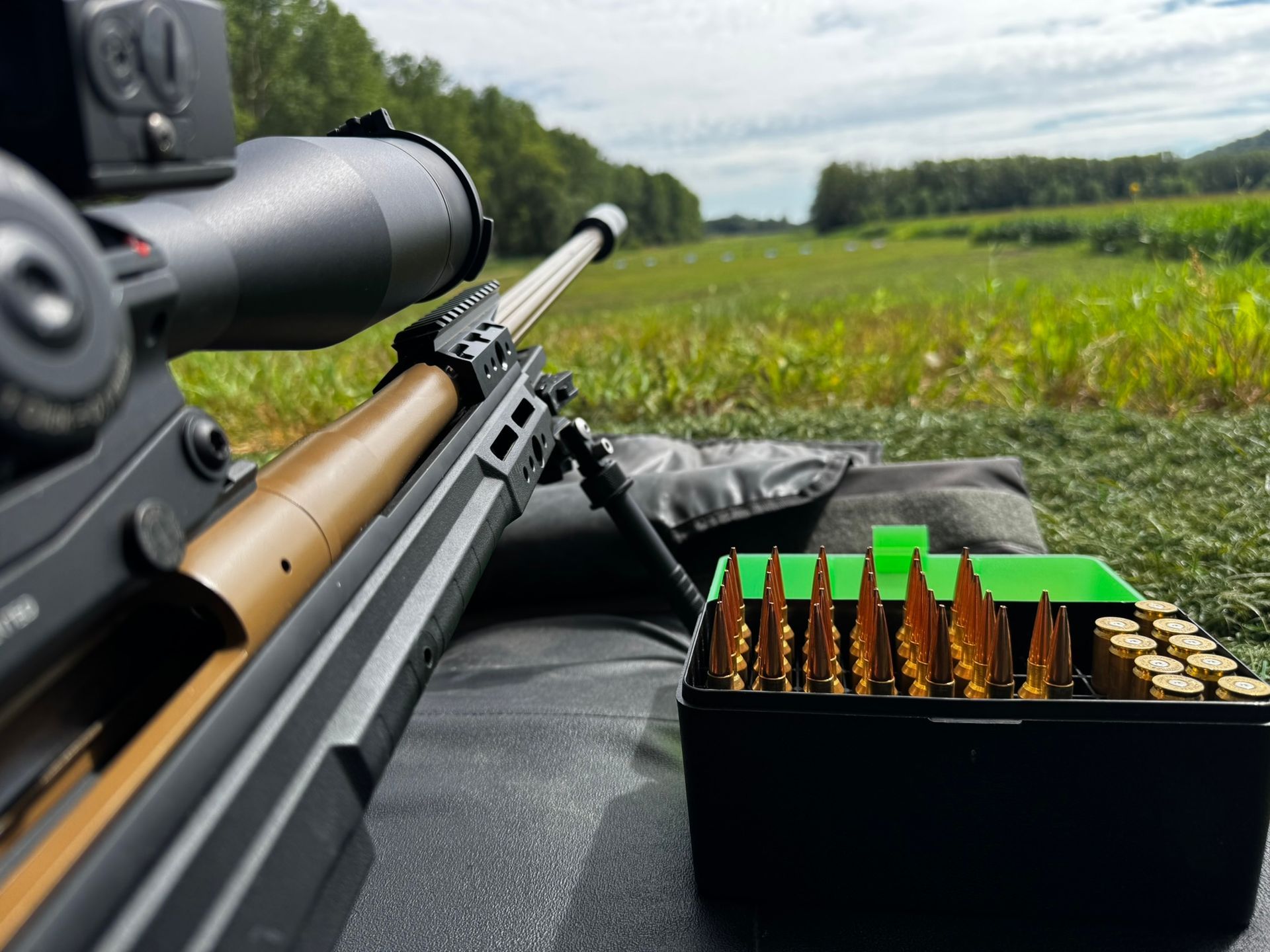 Scharfschützengewehr mit Zielfernrohr und Munition auf einer Matte, auf ein Feld gerichtet.