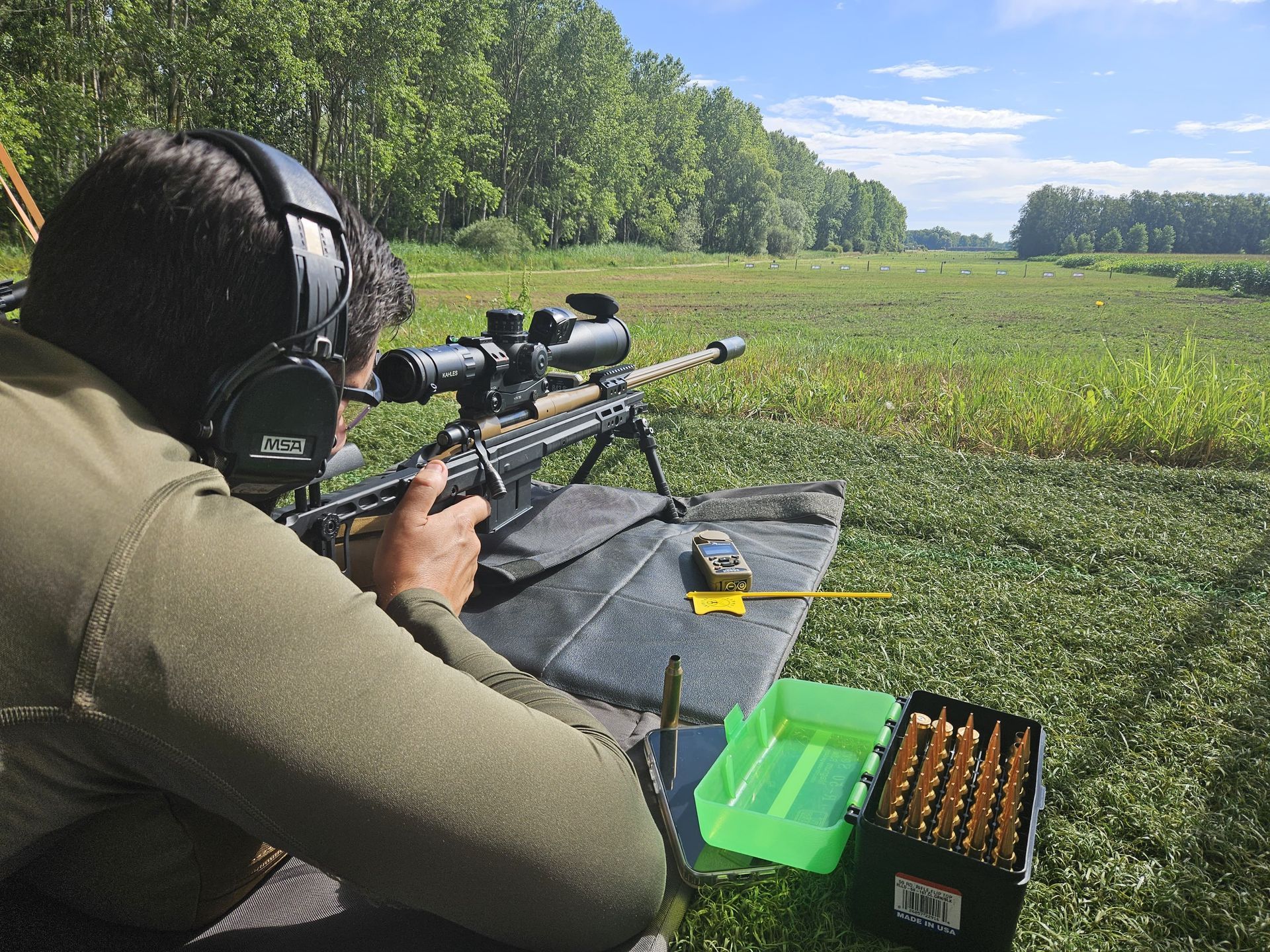 Ein Mann trägt Kopfhörer und zielt mit einem Gewehr mit Zielfernrohr auf ein Feld. Neben ihm steht e