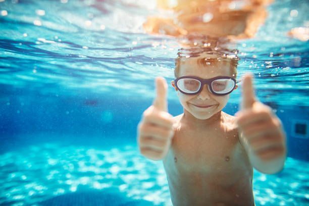A young boy wearing goggles is giving a thumbs up underwater in a swimming pool.