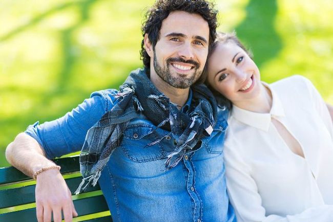 A couple smiles while sitting on a park bench, with the person on the right resting their head on the other's shoulder.