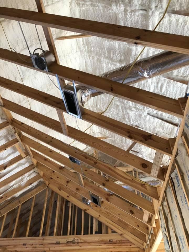 Interior view of unfinished wood roof rafters with spray foam insulation, recessed light housings, and ductwork.