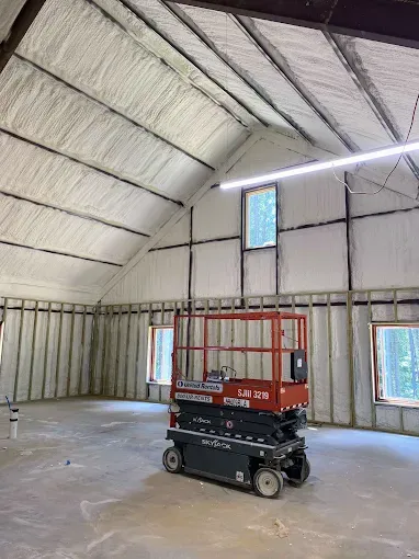 A red scissor lift stands inside an unfinished building with exposed wall framing and spray foam insulation on the roof.