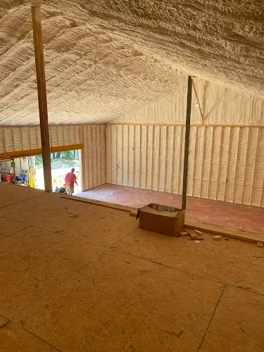 Interior view of an unfinished attic space with spray foam insulation on the walls and ceiling and wooden flooring.