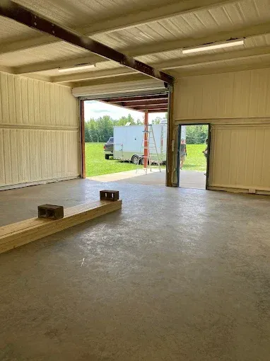 An open bay of a metal workshop with concrete floors, looking out toward a grassy field and a parked trailer.