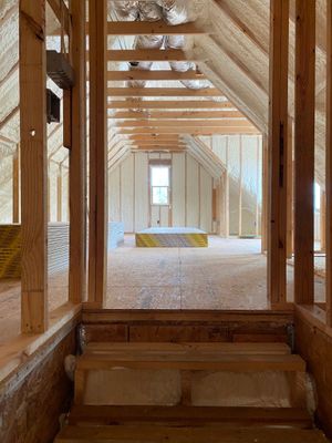 An unfinished attic interior with wooden framing and spray-foam insulation, viewed from the top of a short wooden staircase.