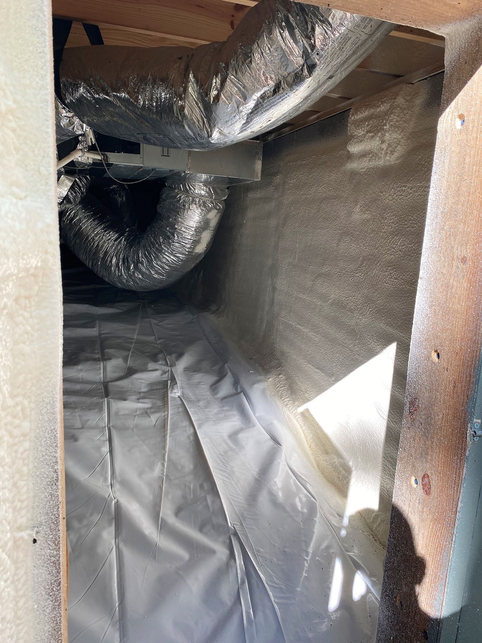 Crawl space with a white vapor barrier on the ground, insulation on the walls, and silver flexible HVAC ductwork above.