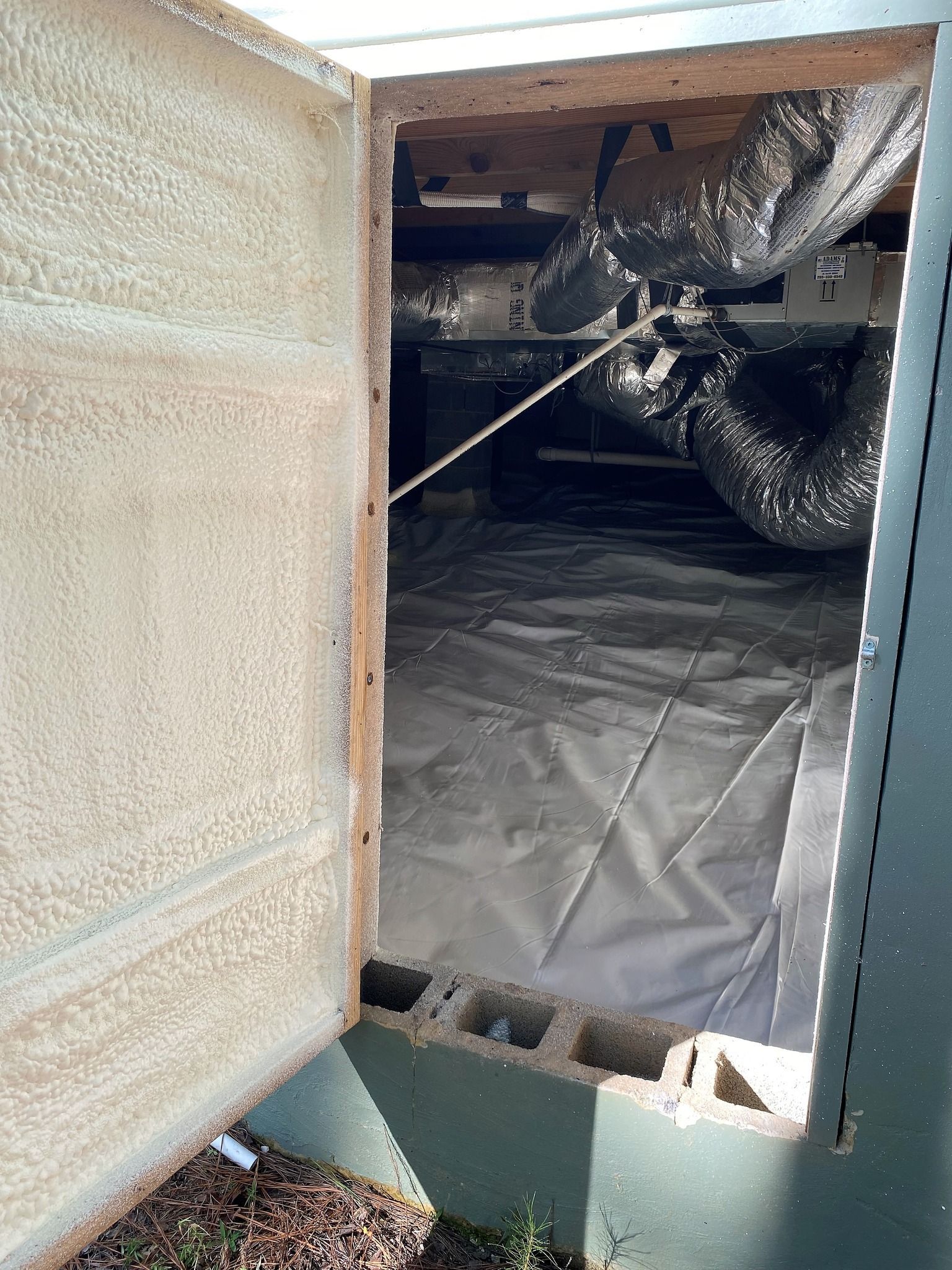 An open crawl space access door reveals insulated silver ductwork and a white vapor barrier covering the ground inside.