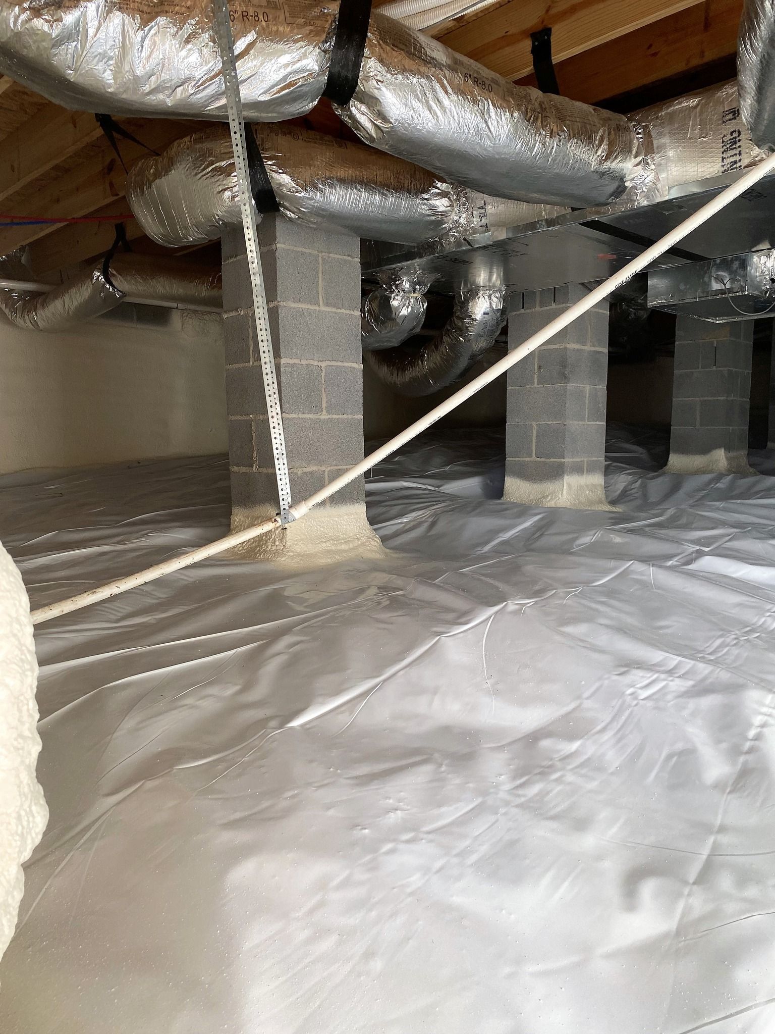 Crawl space with a vapor barrier on the ground, insulated concrete support pillars, and foil-wrapped air ducts overhead.