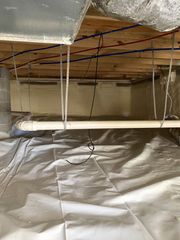 A crawl space with a white floor vapor barrier, overhead wiring, wooden joists, and a horizontal white PVC pipe.