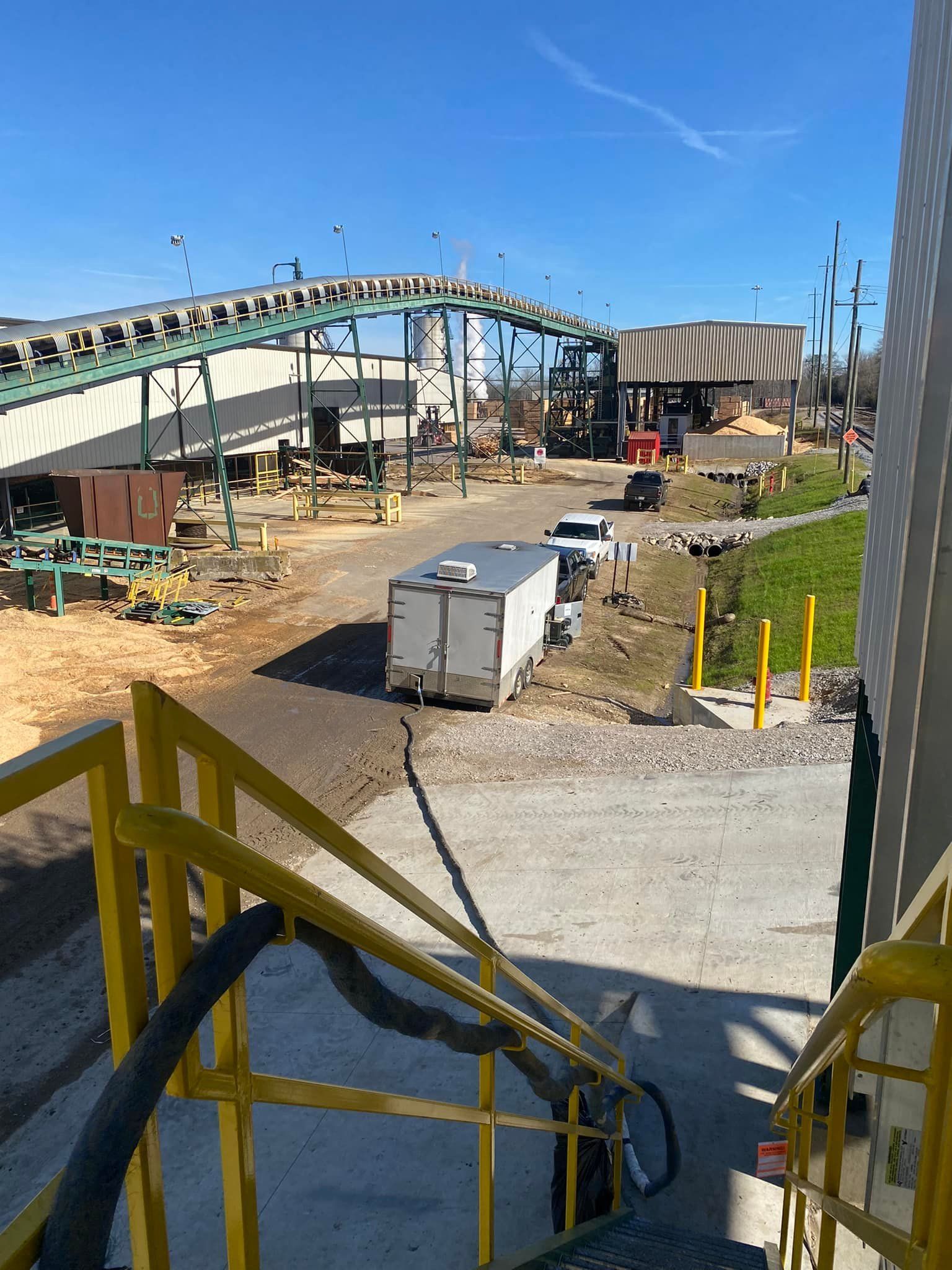 A view looking down yellow stairs at an industrial site featuring a large conveyor belt system and a parked white trailer.