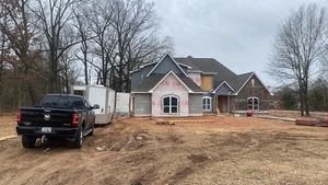 A black pickup truck parked in front of a new house under construction on a dirt lot with bare trees in the background.