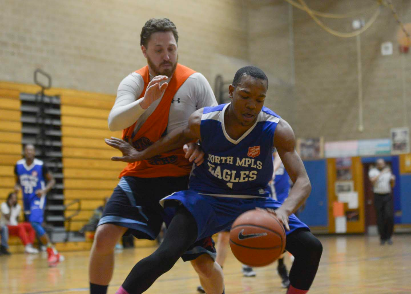 Basketball player in blue jersey dribbling, guarded by a player in orange. Gym setting.