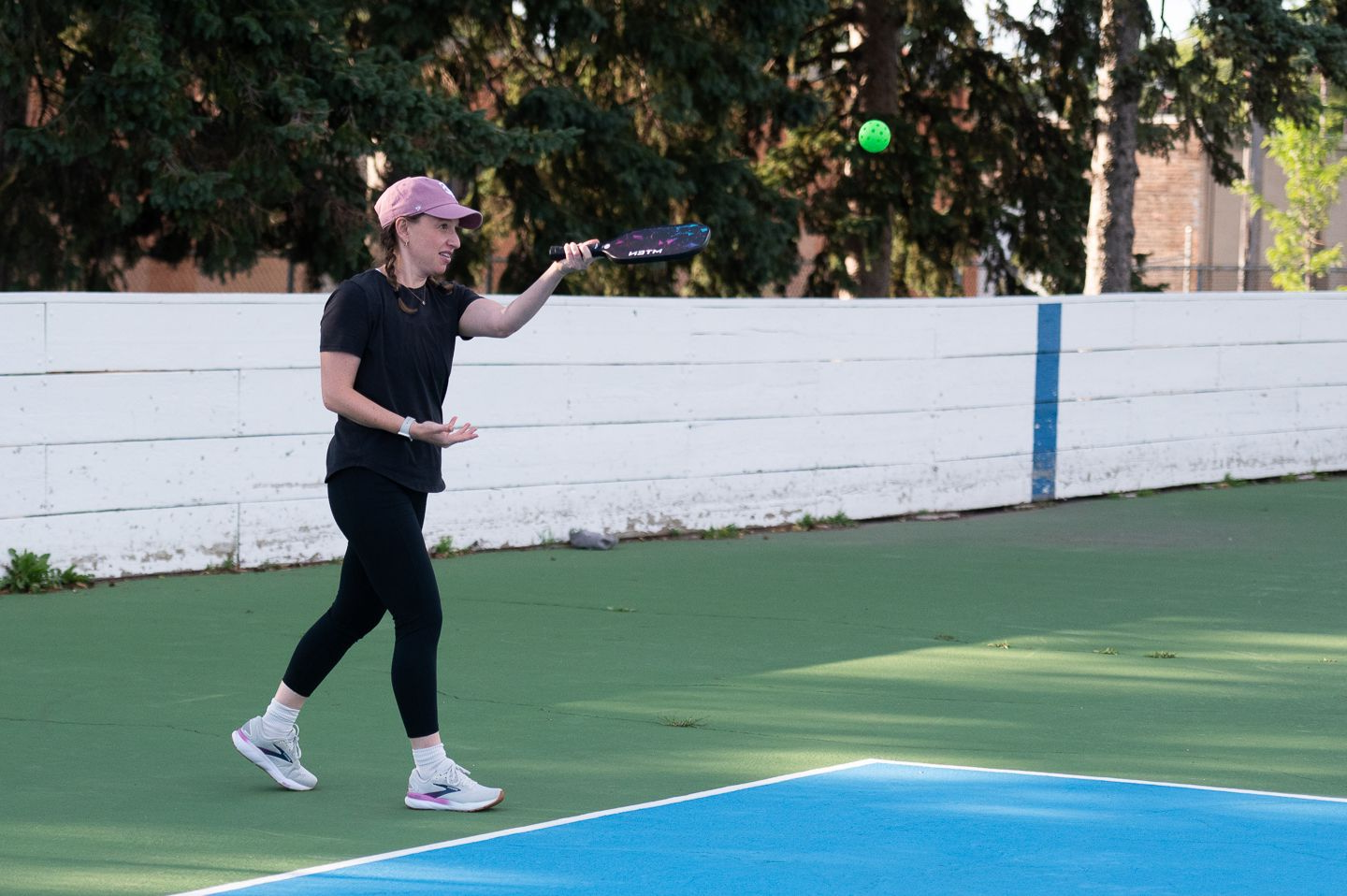 Person on a tennis court holding a racket, reaching for a ball near a blue and green surface.