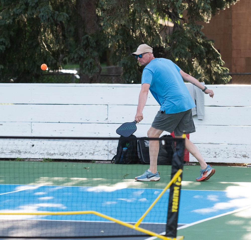Two people playing pickleball on an outdoor court, one in a blue shirt lunging for the ball.
