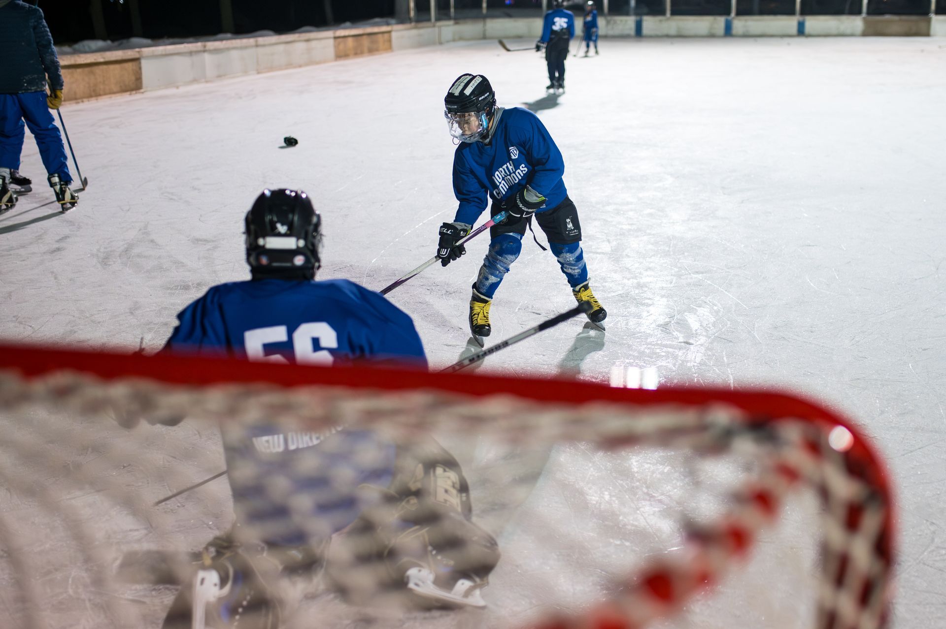 Two hockey players in blue practice on an ice rink while a goalie in yellow stands in the background