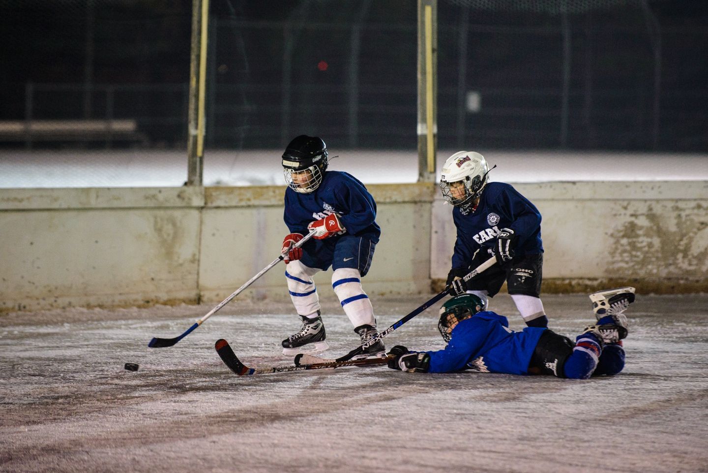 Child in a red vest and black helmet holding a hockey stick on an outdoor ice rink at night