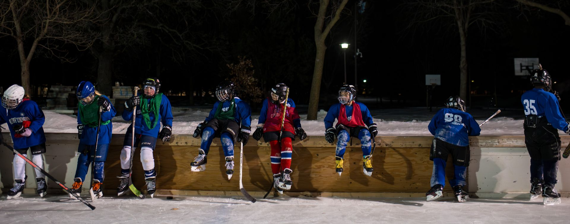Hockey players in blue and red gear line up on an outdoor rink at night, with bare trees in the background.