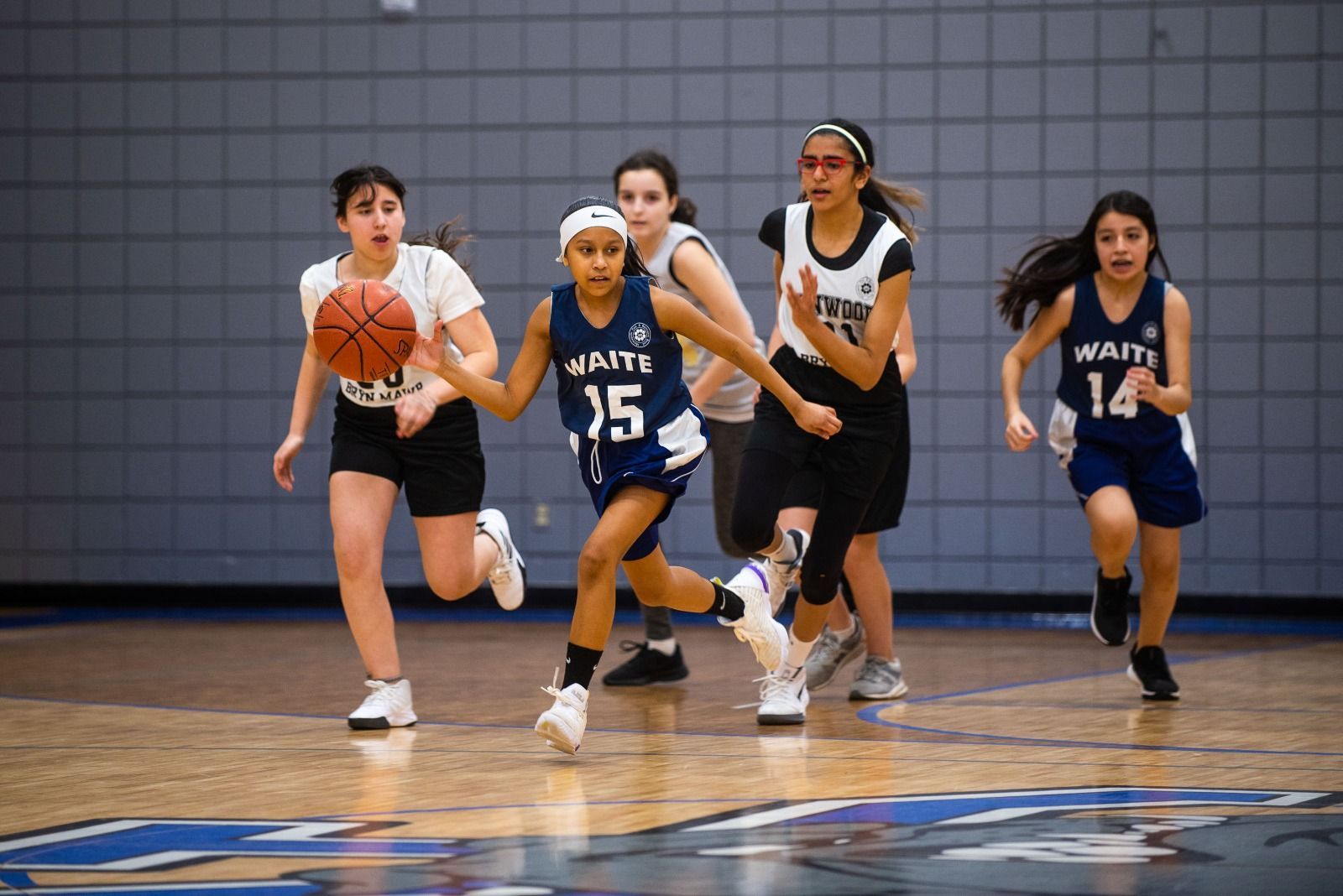 Basketball player in blue jersey dribbling, guarded by a player in orange. Gym setting.