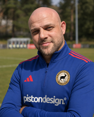 Man in blue sports jacket standing on a grassy field, with a slight smile and a blurred outdoor background