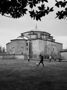Black-and-white view of a stone mosque with a person kicking a soccer ball in the foreground