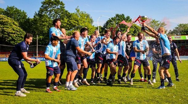 Rugby team posing on a grassy field, celebrating with a coach spraying champagne in bright sunlight