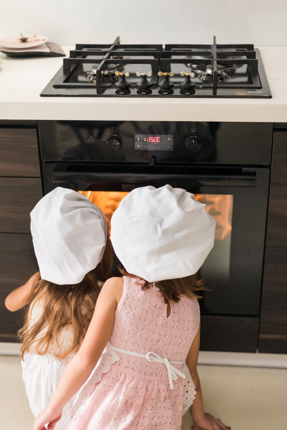 Two children in white chef hats watching a lit oven in a modern kitchen