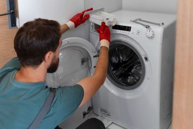Person repairing a front-loading washing machine with a screwdriver in a laundry room