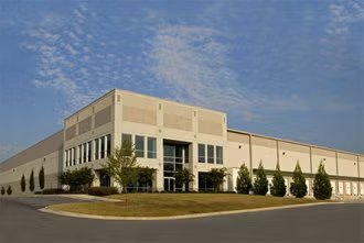 Large beige industrial building with windows and a paved parking lot under a blue sky