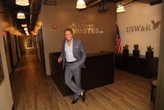 Man in a gray suit standing in a wood-floored office lobby with business signage and an American flag.