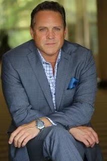 Man in a gray suit sitting with arms crossed, wearing a blue shirt and pocket square.